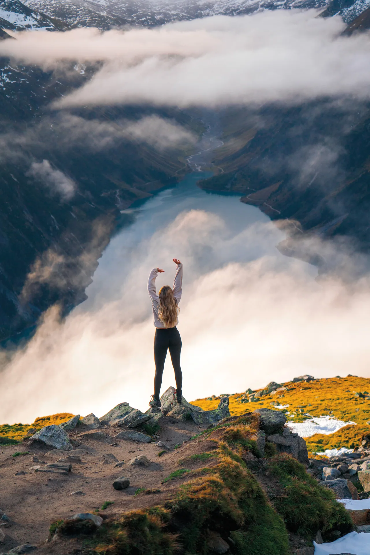 Frau mit erhobenen Armen auf einem Felsen über einem wolkenverhangenen Bergsee